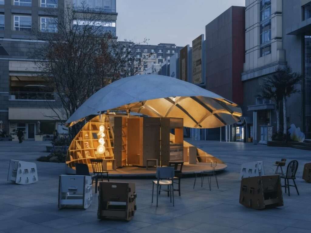 An exterior view of the kinetic "Book Shell" pavilion in Beijing at dusk, semi-open and glowing with warm internal light. People are scattered in the plaza on movable cardboard seating.