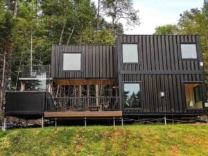 Exterior view of Fairmont Villa, a two-story modern container home with matte black finish and large windows in a forest setting.