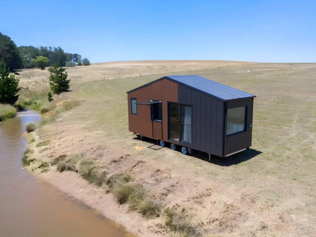 Exterior view of Audrey tiny home by CozyCo featuring wood-textured panels and corrugated aluminum cladding in a rural landscape.