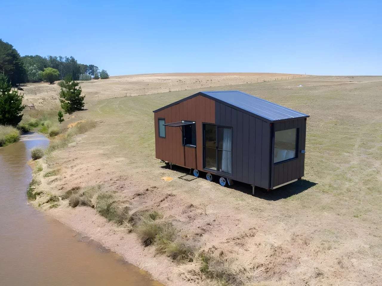 Exterior view of Audrey tiny home by CozyCo featuring wood-textured panels and corrugated aluminum cladding in a rural landscape.