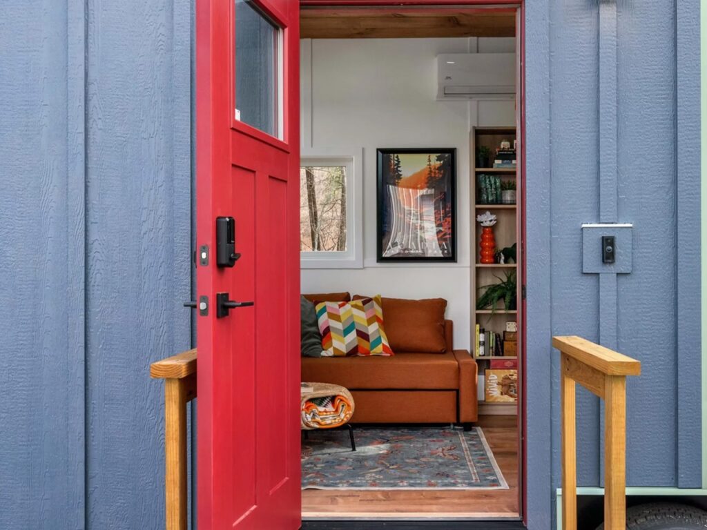 A vibrant red front door of a tiny house opening into a modern living room with a brown sofa and colorful decor.