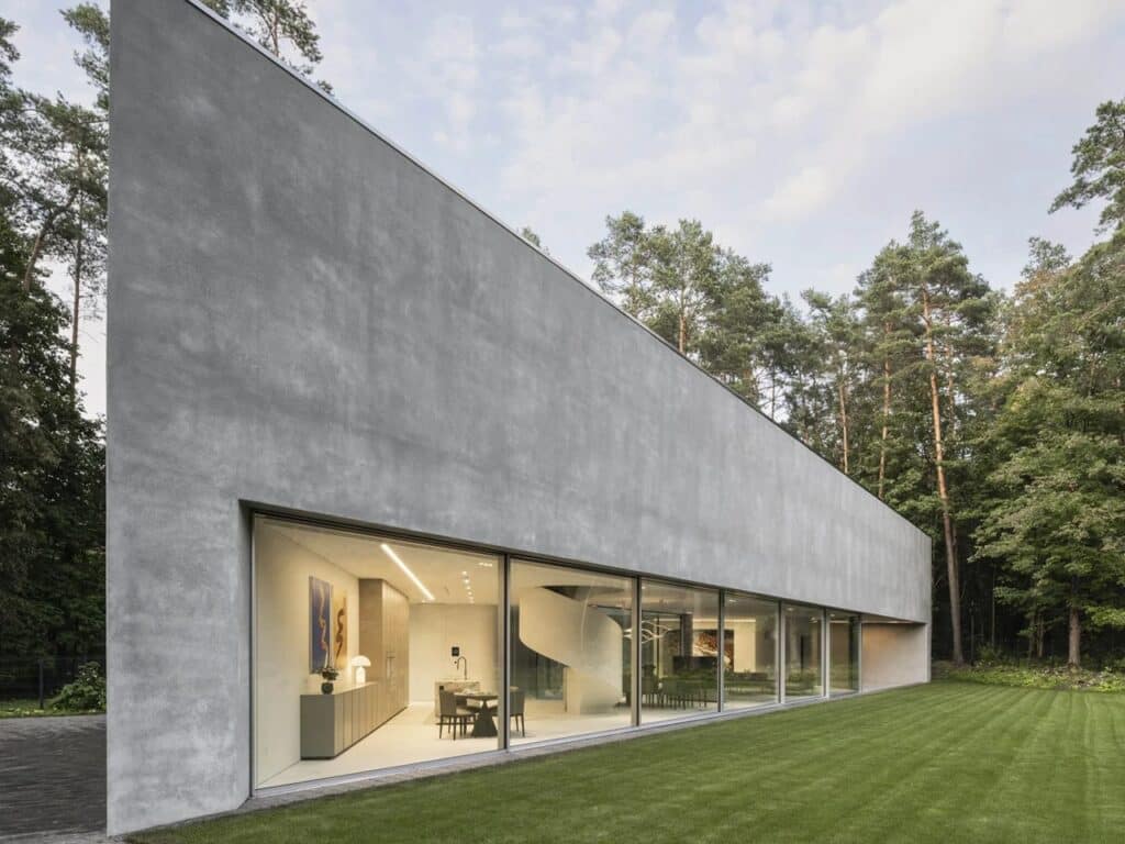 Side view of Trim House showing a long minimalist concrete facade with expansive floor-to-ceiling glass windows in a forest setting.