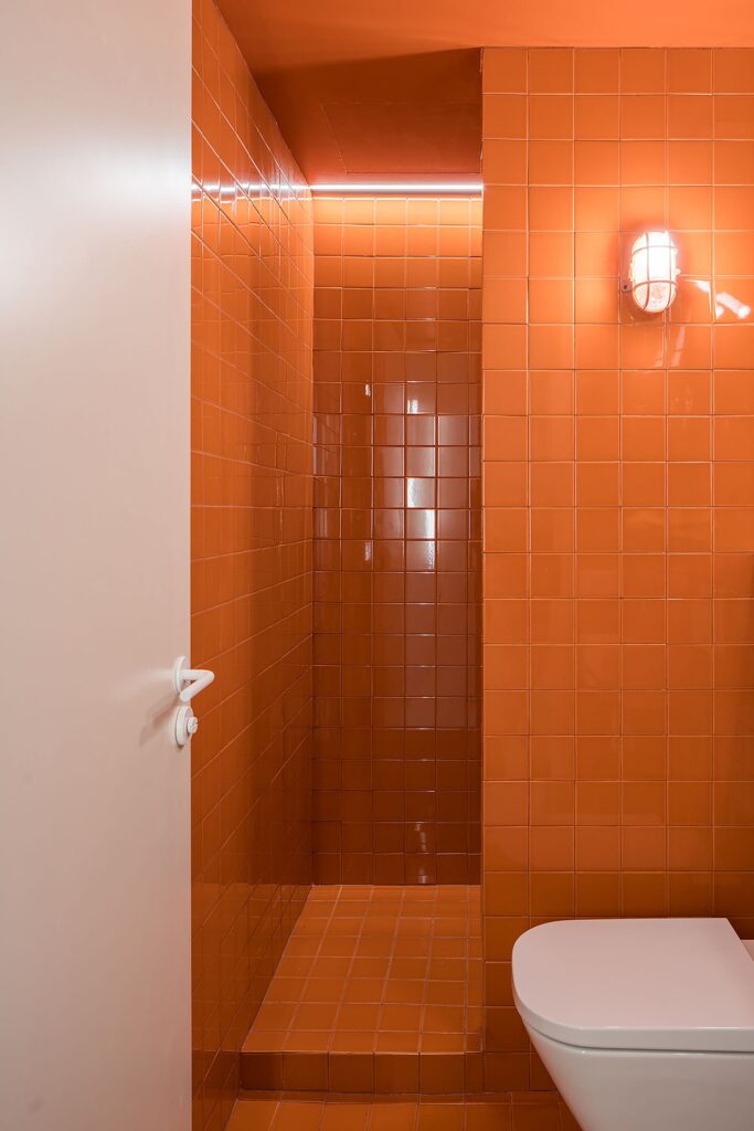 A modern bathroom interior fully clad in vibrant orange ceramic tiles with industrial-style bulkhead lighting.