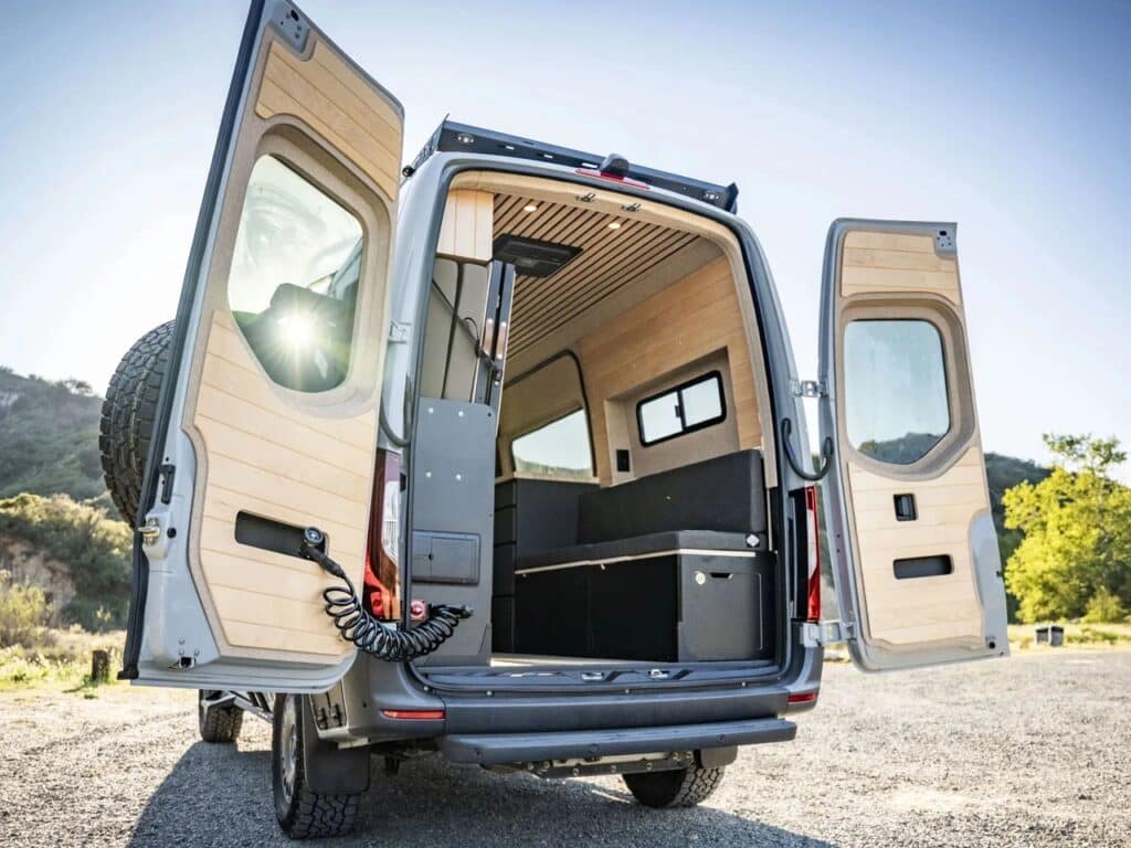 Rear view of the camper van with open back doors, revealing wood-paneled interior walls and an L-shaped seating area.