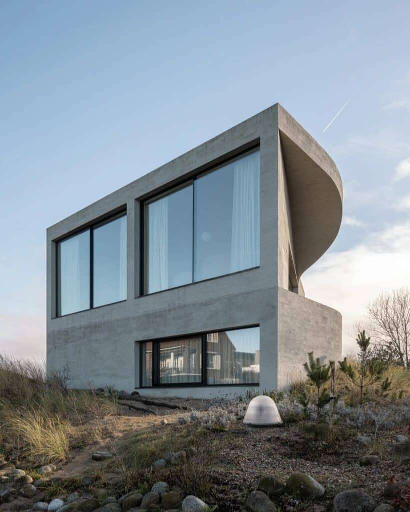 Exterior of a modern concrete villa with large glass windows integrated into a coastal dune landscape.