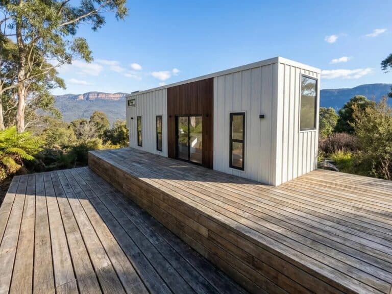 Exterior view of Onda tiny house on wheels featuring white corrugated steel cladding and a large wooden deck in a forest setting.