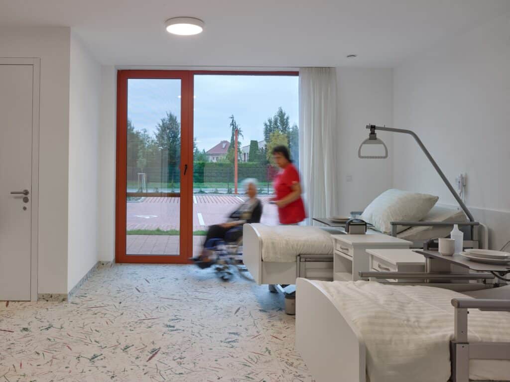 Interior of a resident's room featuring a medical bed, large window with a view, and nature-patterned flooring.