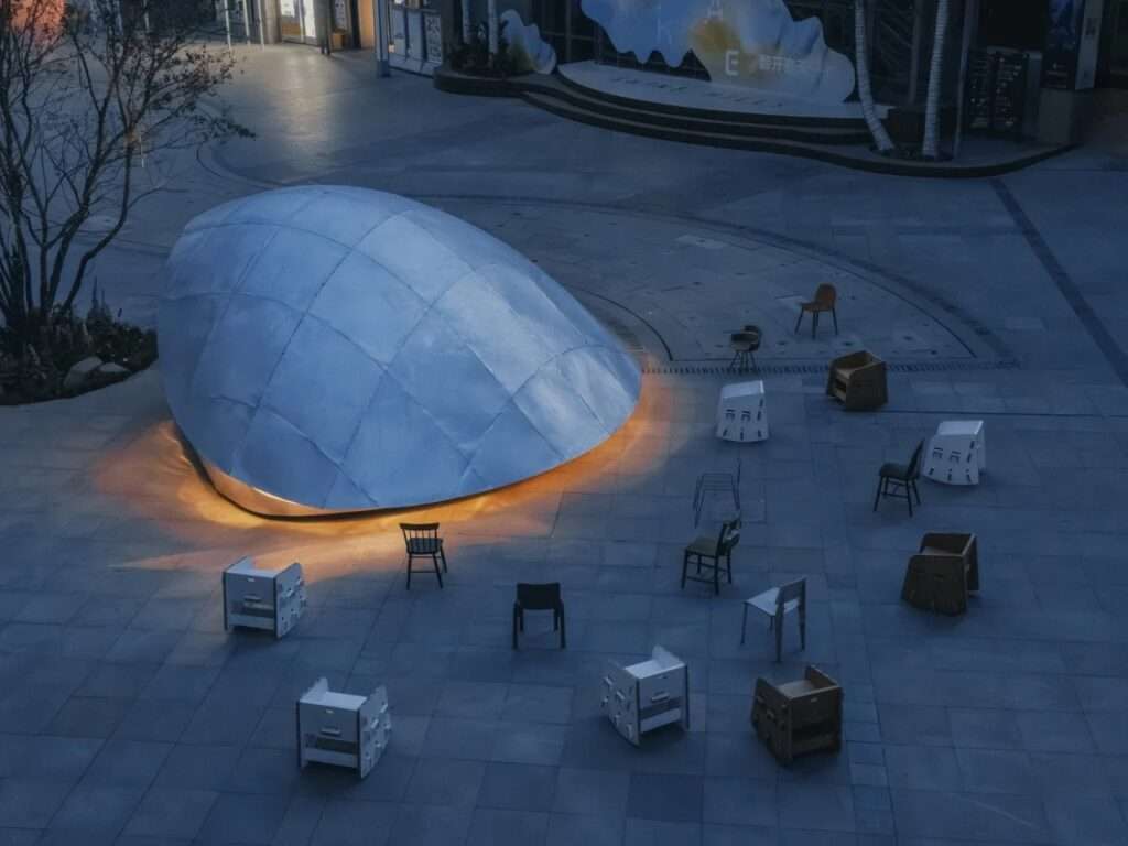 An aerial high-angle view of the "Book Shell" pavilion fully closed into its sleek metallic shell form at dusk, casting a warm glow at its base in an empty plaza.
