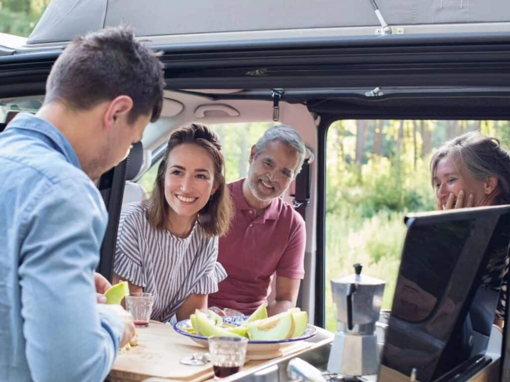 A group of people smiling and enjoying a meal inside the Tonke Baycamper kitchen area with a view of the outdoors.