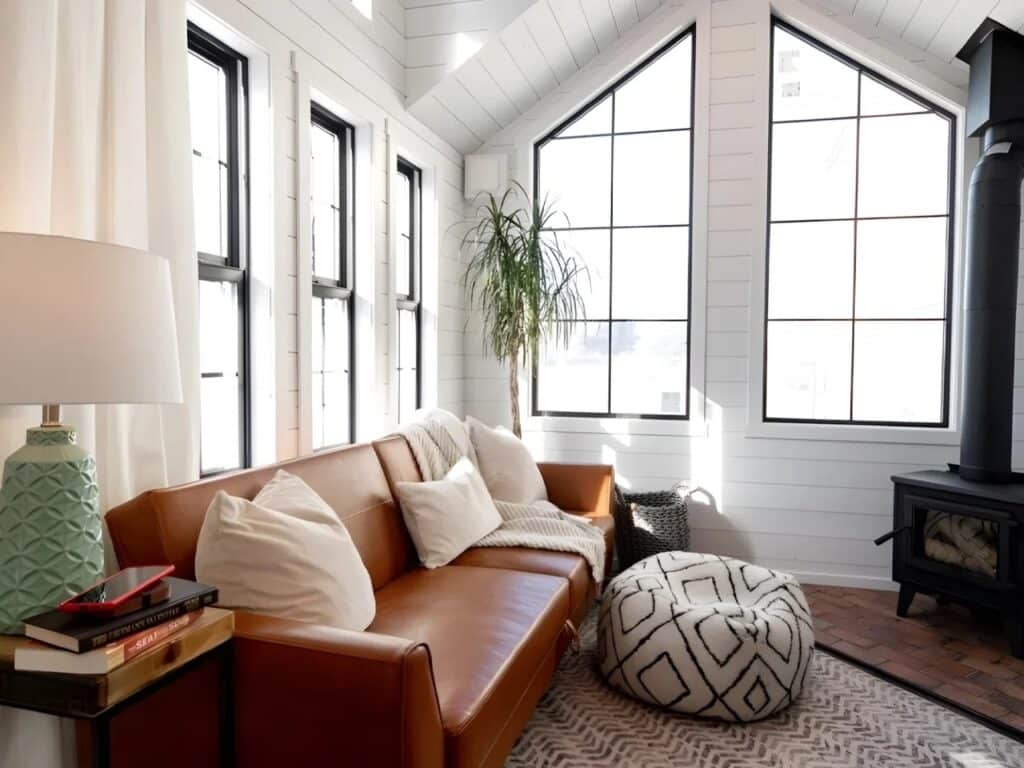 Bright interior living room of Daphne tiny house with a leather sofa, wood-burning stove, and large geometric windows.