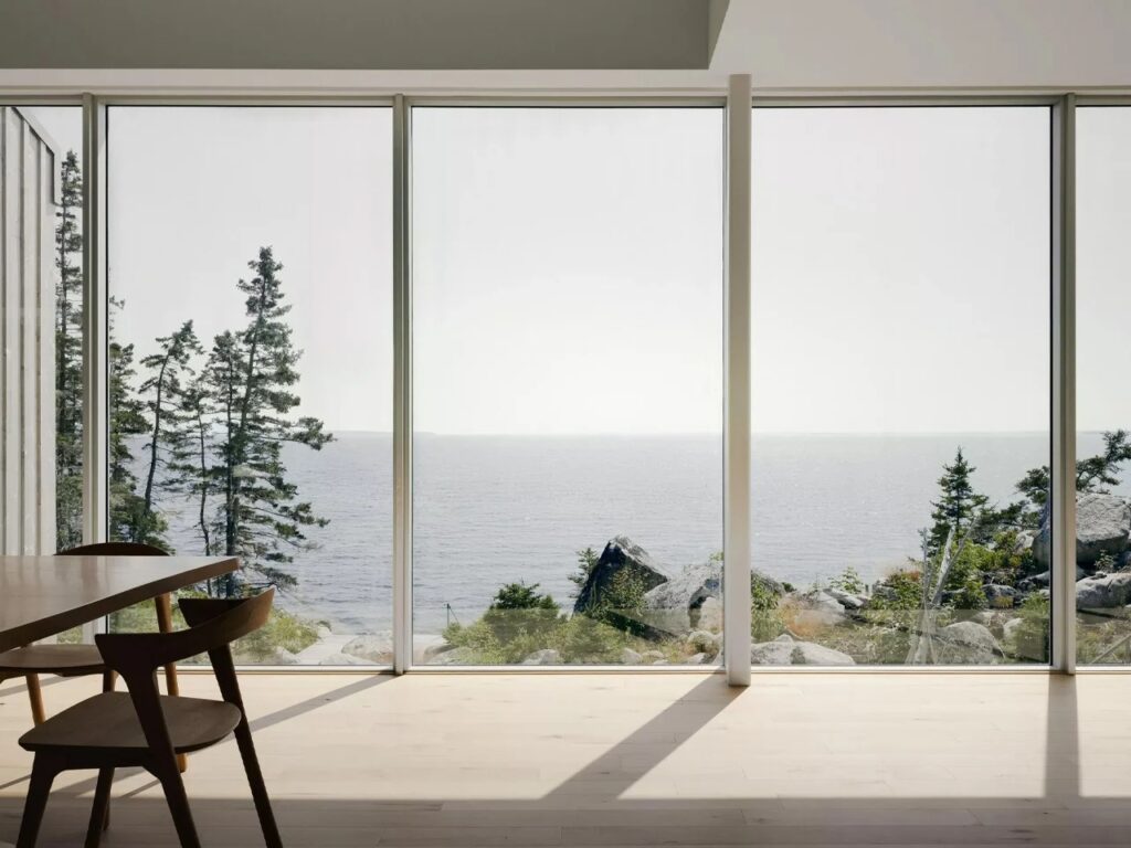 Interior view of East River Residence dining area with large floor-to-ceiling glass windows overlooking the Atlantic Ocean and coastal trees.