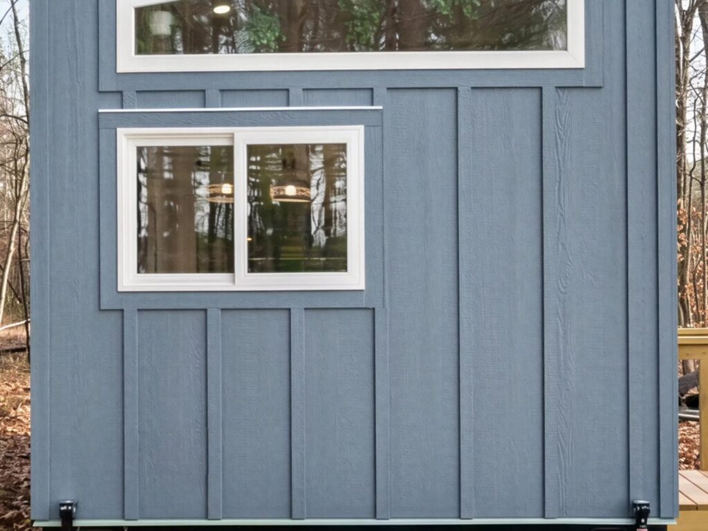 Close-up of blue board and batten siding on a tiny house with white-trimmed windows reflecting the surrounding forest.