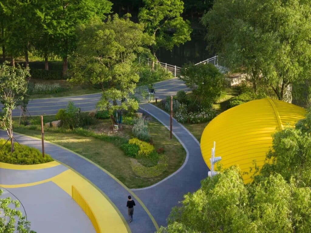 Winding grey walking paths with yellow borders passing through lush greenery and a bright yellow curved structure in Kunshan.