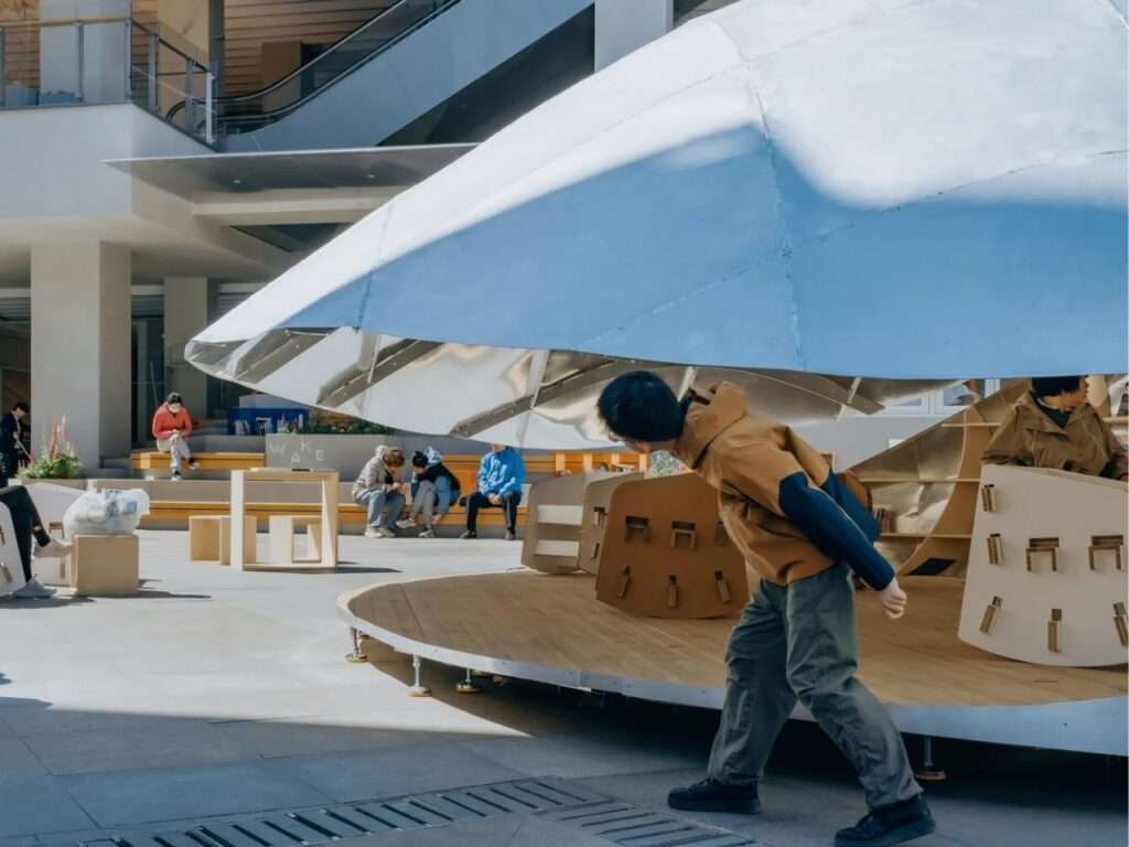 A side-profile photograph of a user interacting with the "Book Shell" pavilion. A man in a brown jacket leans forward to browse bookshelves under the partially opened metallic roof.