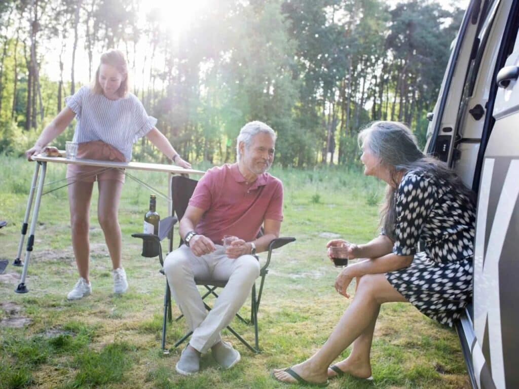 A family relaxing outside the Tonke camper van in a sunny forest clearing, featuring outdoor seating and a portable table.
