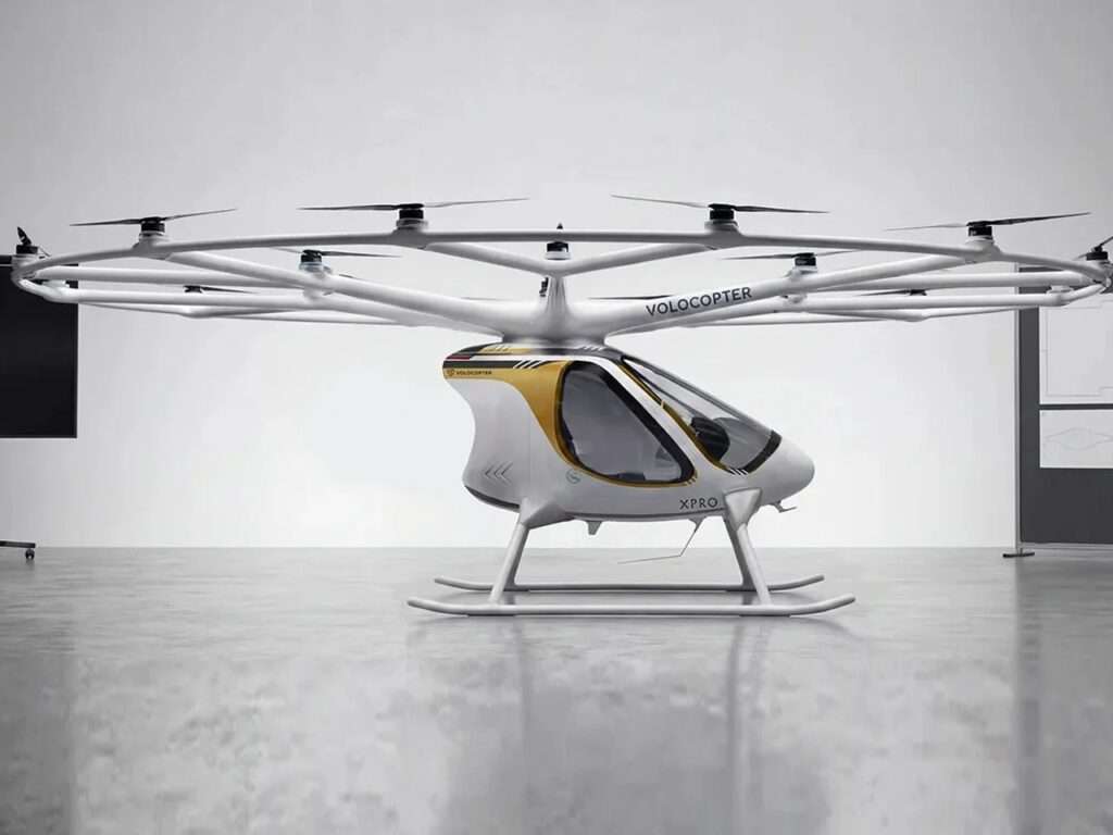 A full front three-quarter view of the white Volocopter VoloXPro eVTOL aircraft, showing the cabin with its teal nose cone and the extensive 18-rotor overhead ring structure.