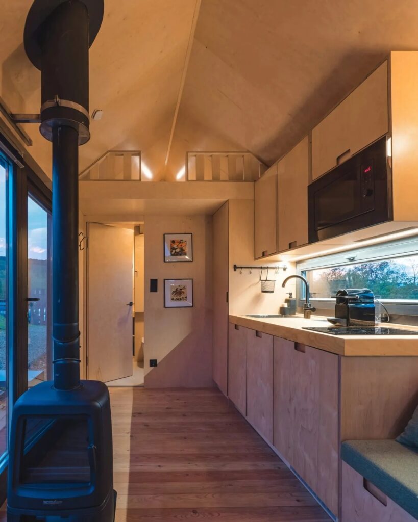 Interior of NestOff tiny house showing birch plywood kitchen cabinets, a black wood-burning stove, and a view through the door.