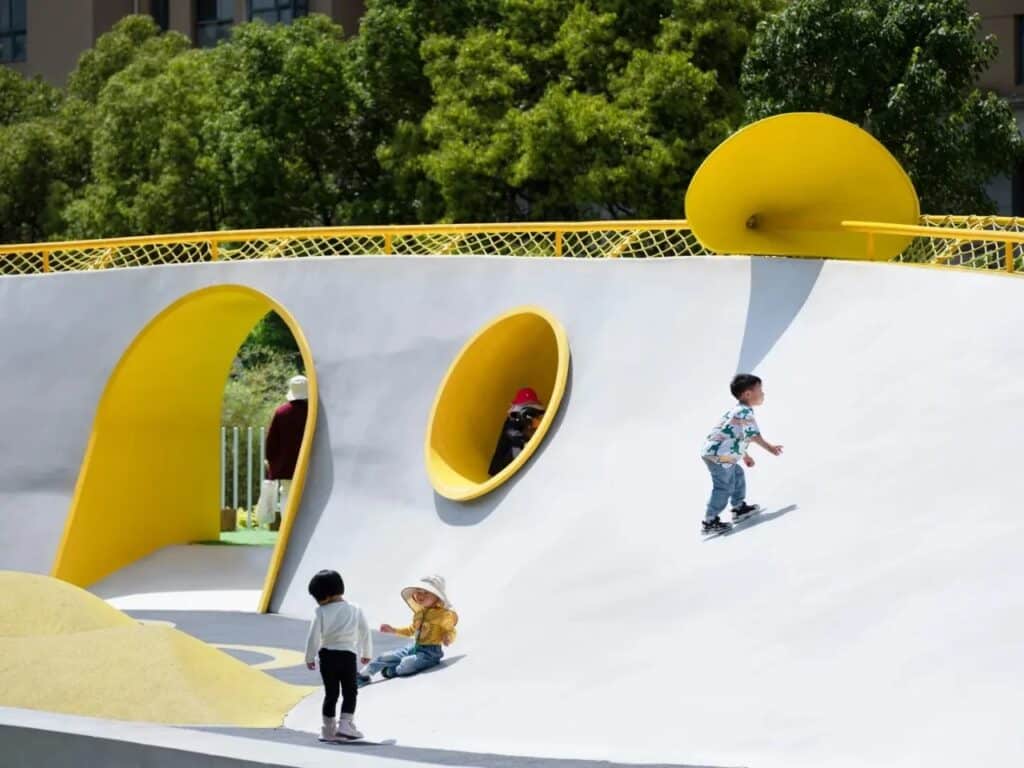 Children playing on a white sculptural wall with bright yellow circular tunnels and trumpet-shaped installations.