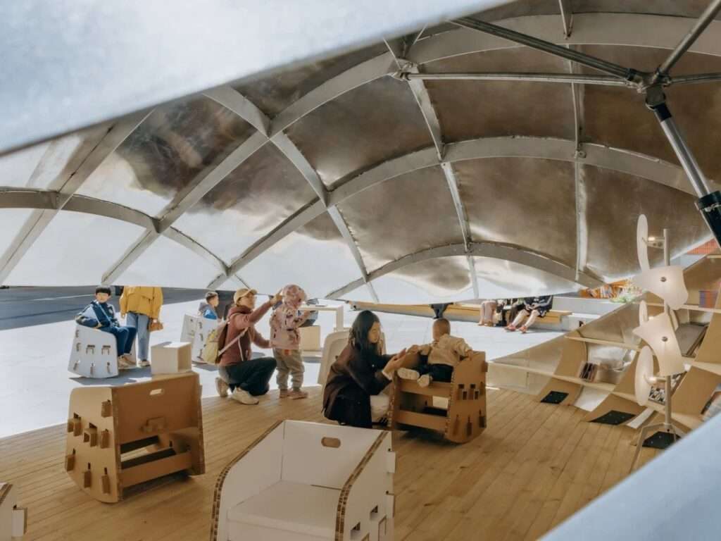 An interior viewpoint from inside the "Book Shell" pavilion looking out. A mother and child interact on the wooden deck while the curved metallic structure is visible overhead.