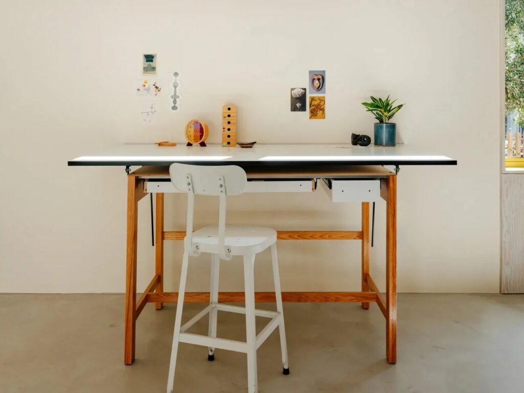Close-up of a white drafting table and stool against a minimalist gallery wall in an architectural studio.