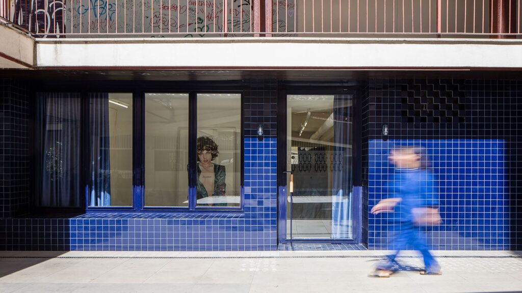 Frontal view of the blue ceramic facade with large glass windows and a blurred pedestrian walking past.
