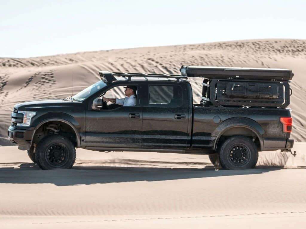 Side view of a black truck with a closed Altus hardwall tent driving through desert dunes.