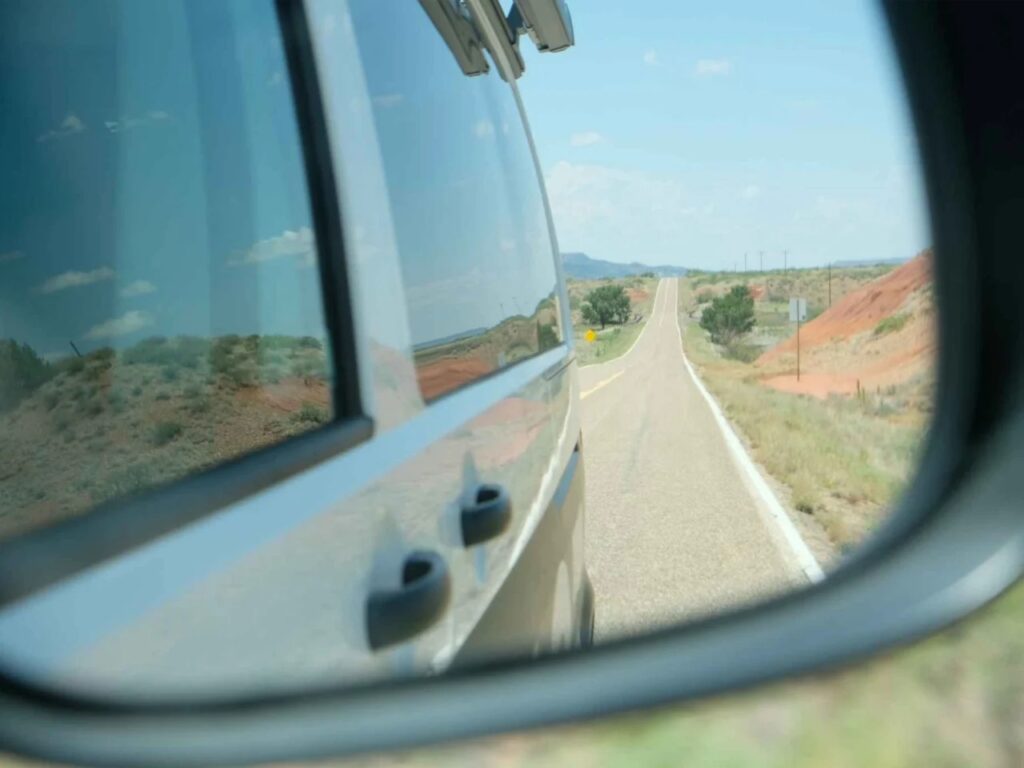 View through a side mirror of a camper van driving on an open desert highway under a clear blue sky.