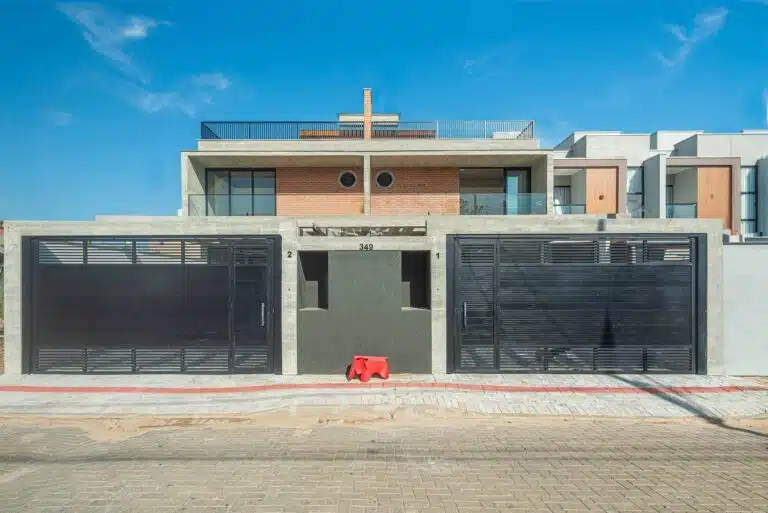 Modern twin house facade with exposed concrete, brickwork, and black metal gates under a blue sky.