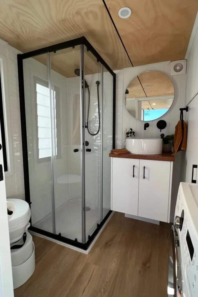 Compact modern bathroom in a tiny home featuring a glass shower, round mirror, and white vanity.
