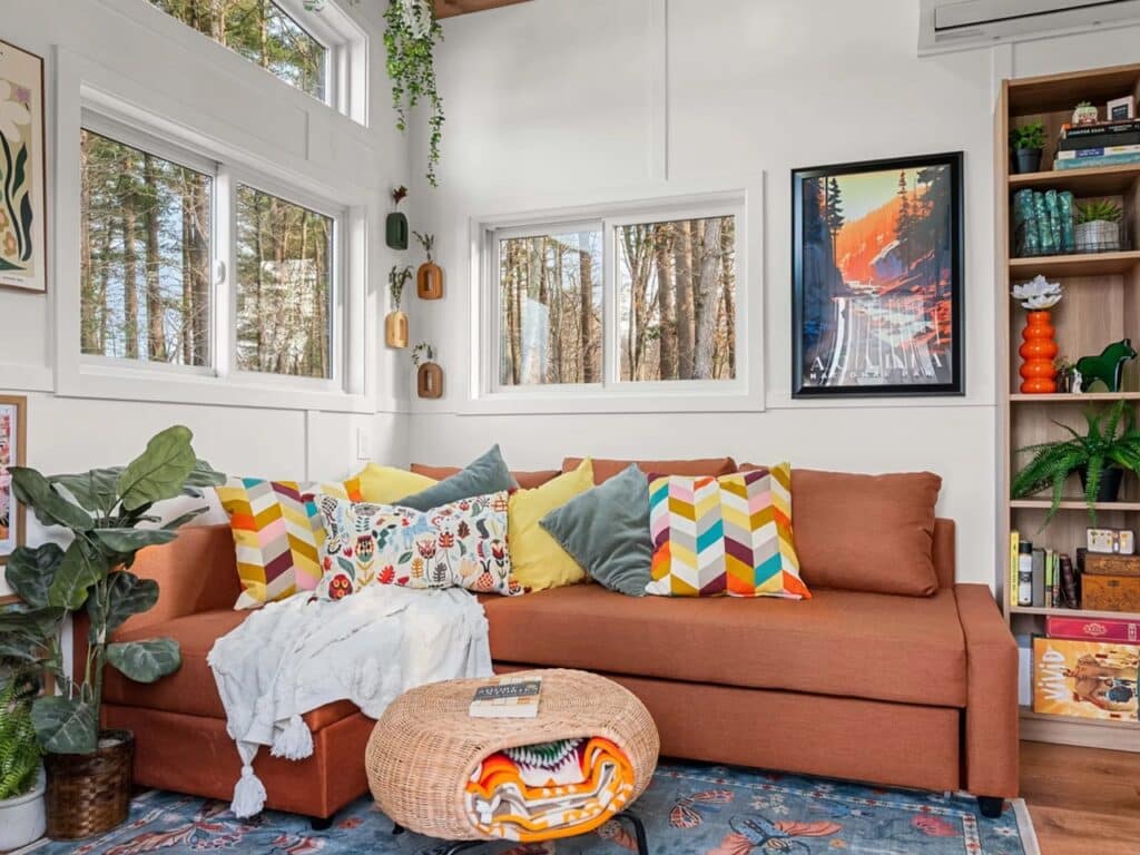 A cozy living room in a tiny house with an orange-brown sofa, geometric pillows, and a tall wooden bookshelf.