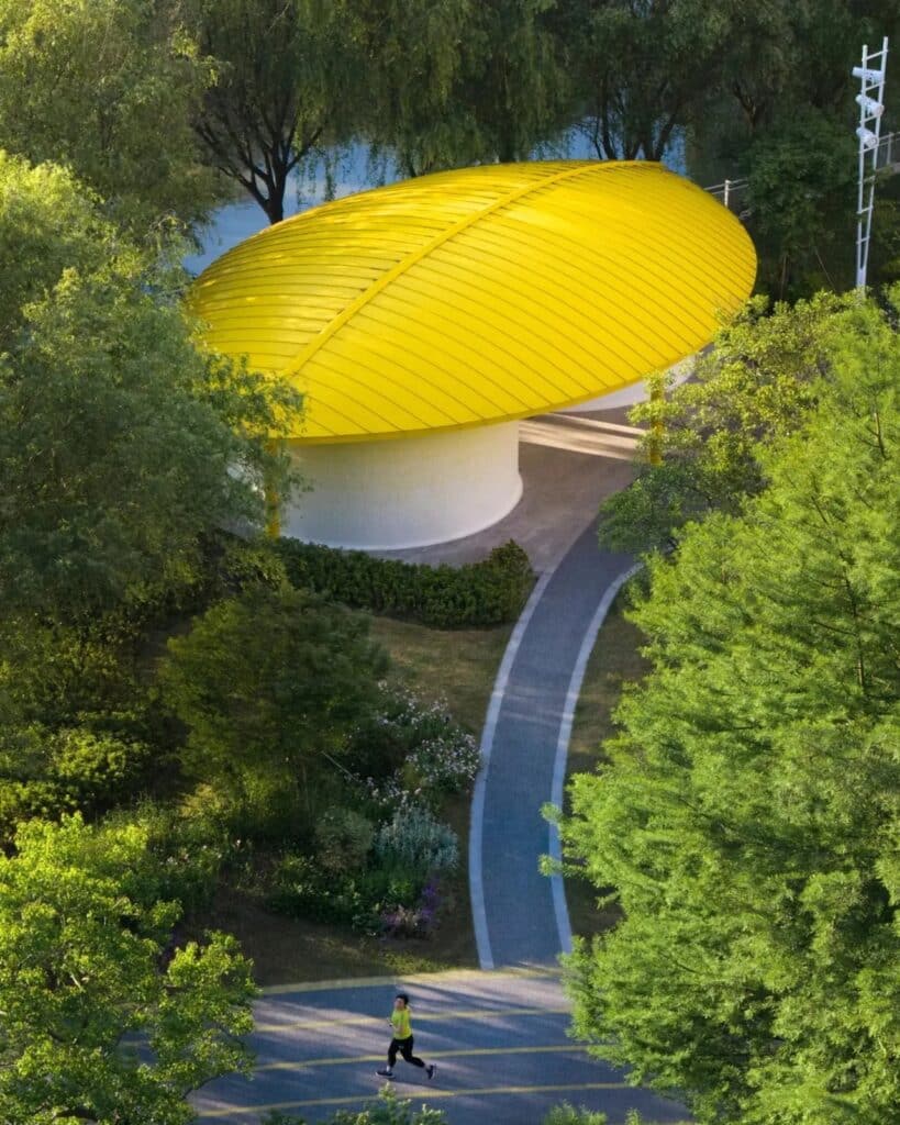 A bright yellow oval pavilion roof seen through a frame of green trees with a jogger on a path below.