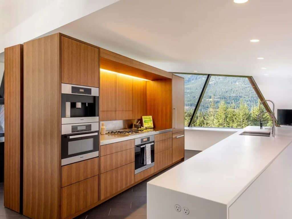 Minimalist kitchen in Hadaway House with warm wood cabinetry, integrated appliances, and a large white island facing a scenic window.