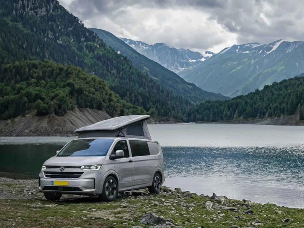 Silver Tonke Baycamper parked by a serene mountain lake with snow-capped peaks in the background.