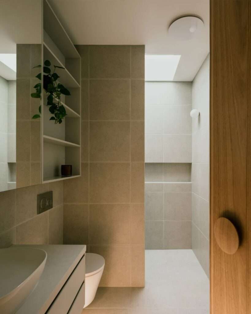Modern minimalist bathroom with a skylight providing natural light over beige tiles and a wooden door detail.