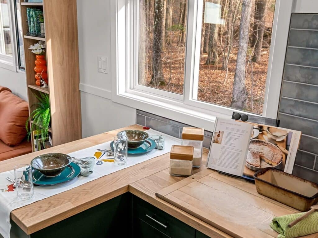 A wooden kitchen countertop in a tiny house with a view of the woods through a large window, featuring a breakfast bar setup.