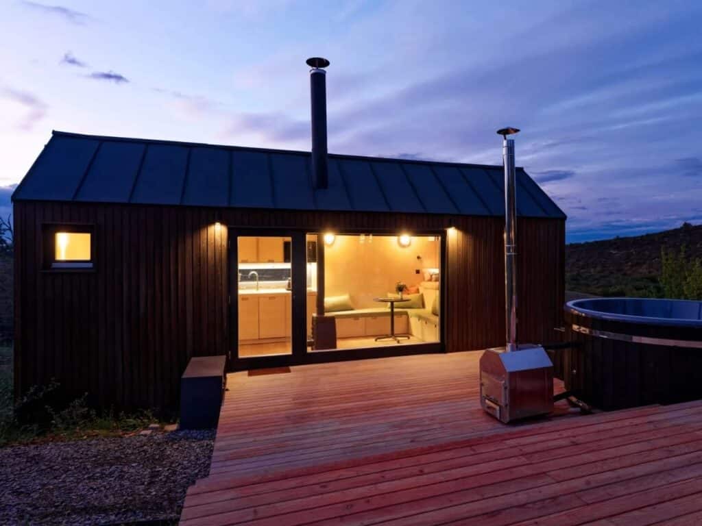 Side view of the NestOff dark wooden cabin at dusk, showing the illuminated interior and the multi-level terrace.