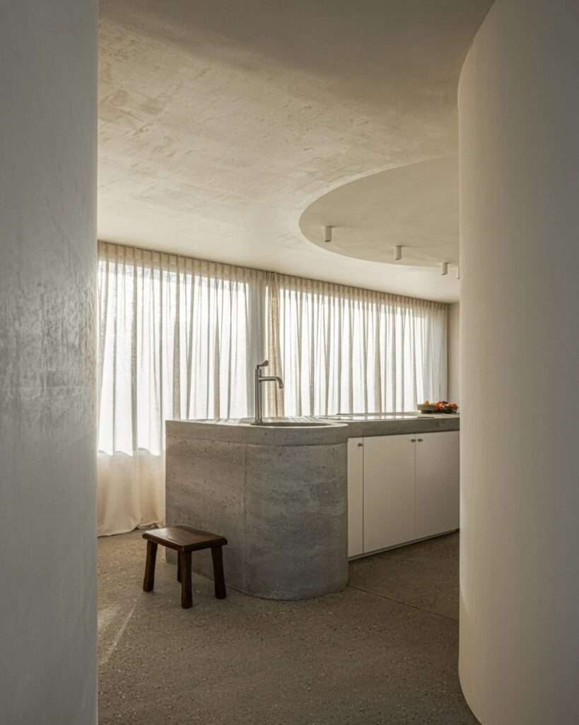 Interior of a minimalist kitchen with a custom curved concrete island and soft natural lighting.