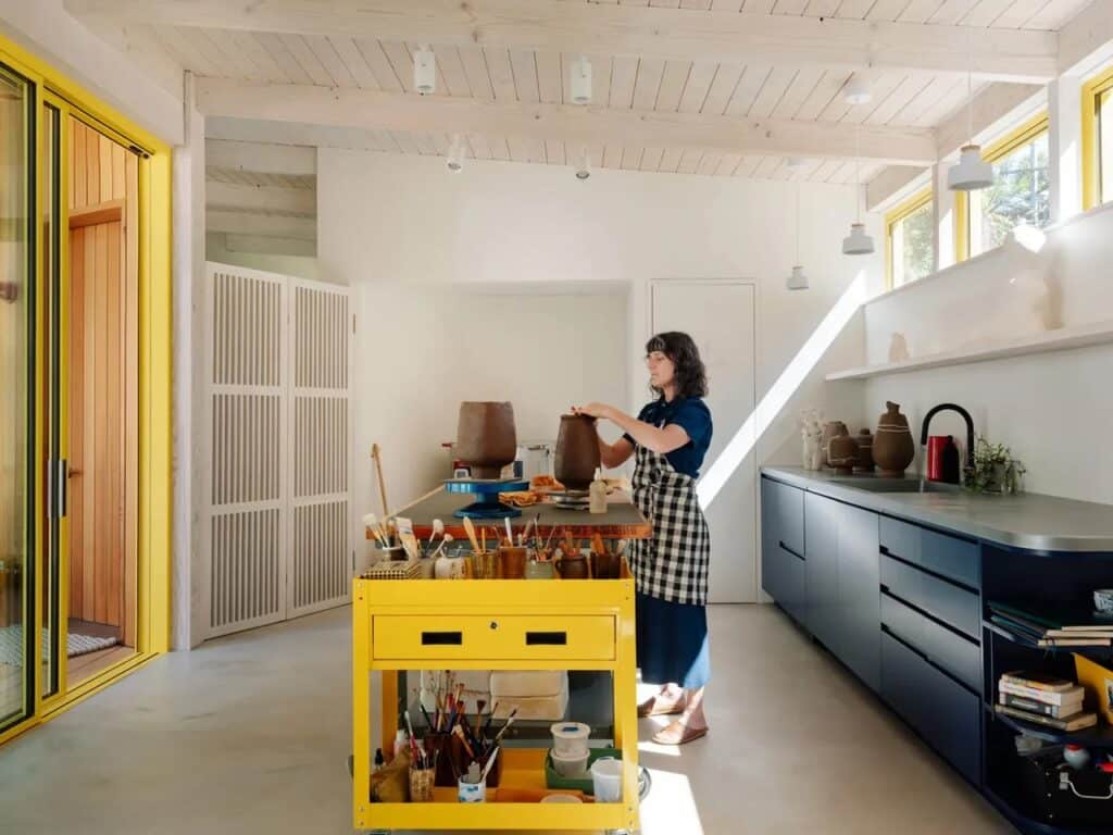 Interior of a pottery workshop with a yellow tool cart, blue kitchen cabinets, and high clerestory windows.