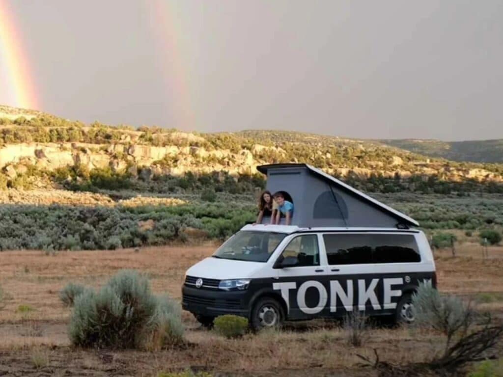 Two children looking out from the pop-up roof tent of a Tonke camper van under a double rainbow in a desert landscape.
