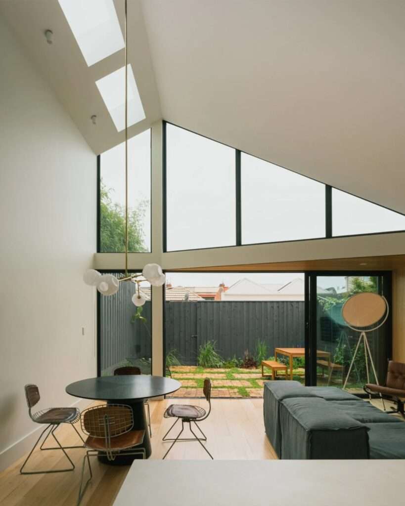 Open plan living and dining room with a view of a brick patio and black timber fence under a dramatically pitched ceiling.