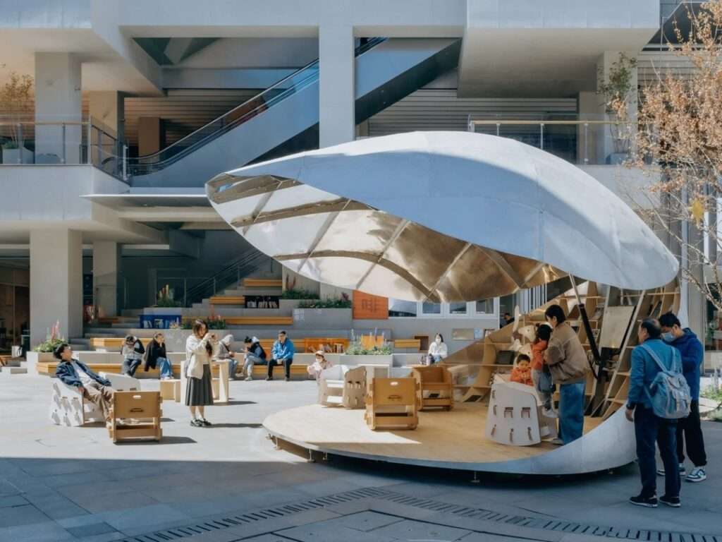 An exterior daytime photograph of the semi-open "Book Shell" pavilion in a bustling commercial plaza with families and children sitting on its wooden deck.