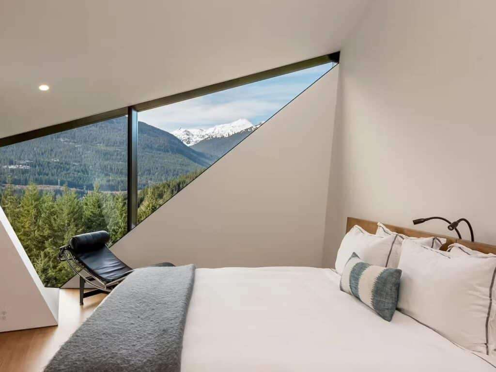 Modern bedroom in Hadaway House with a white bed, a black lounger, and a large triangular window framing a snow-capped mountain.