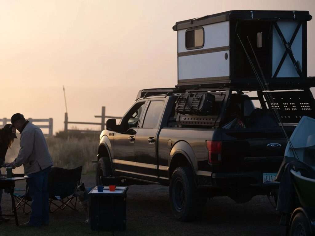 People preparing a meal at a campsite next to a truck with an Altus rooftop cabin at dusk.