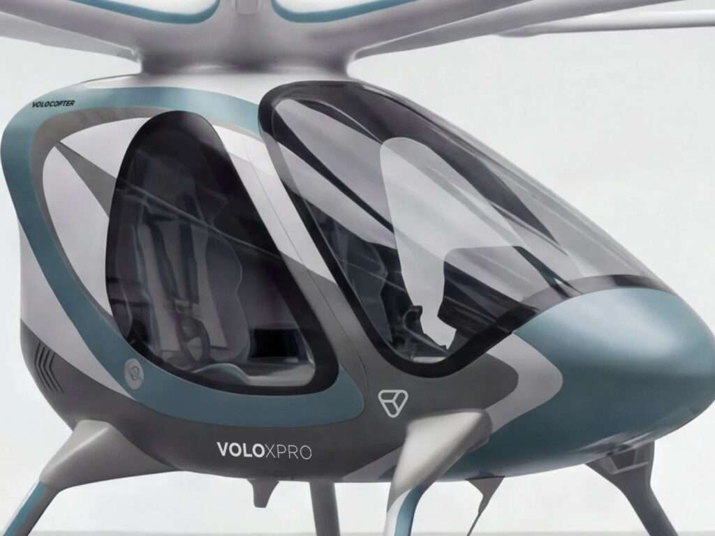 A medium front three-quarter close-up view of the Volocopter VoloXPro cabin, emphasizing the two-seat configuration and the panoramic glass canopy with a teal-blue nose.