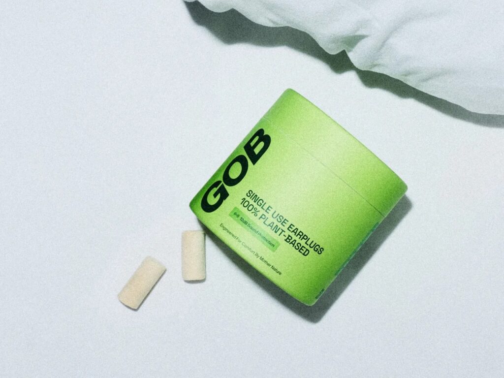 Flat lay view of GOB plant-based earplugs next to their green paperboard container on a white textured surface.
