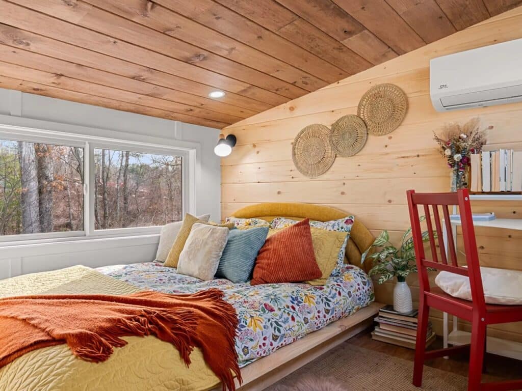 A loft bedroom in a tiny house with a bed, wooden walls, a small desk with a red chair, and a ceiling-mounted AC unit.