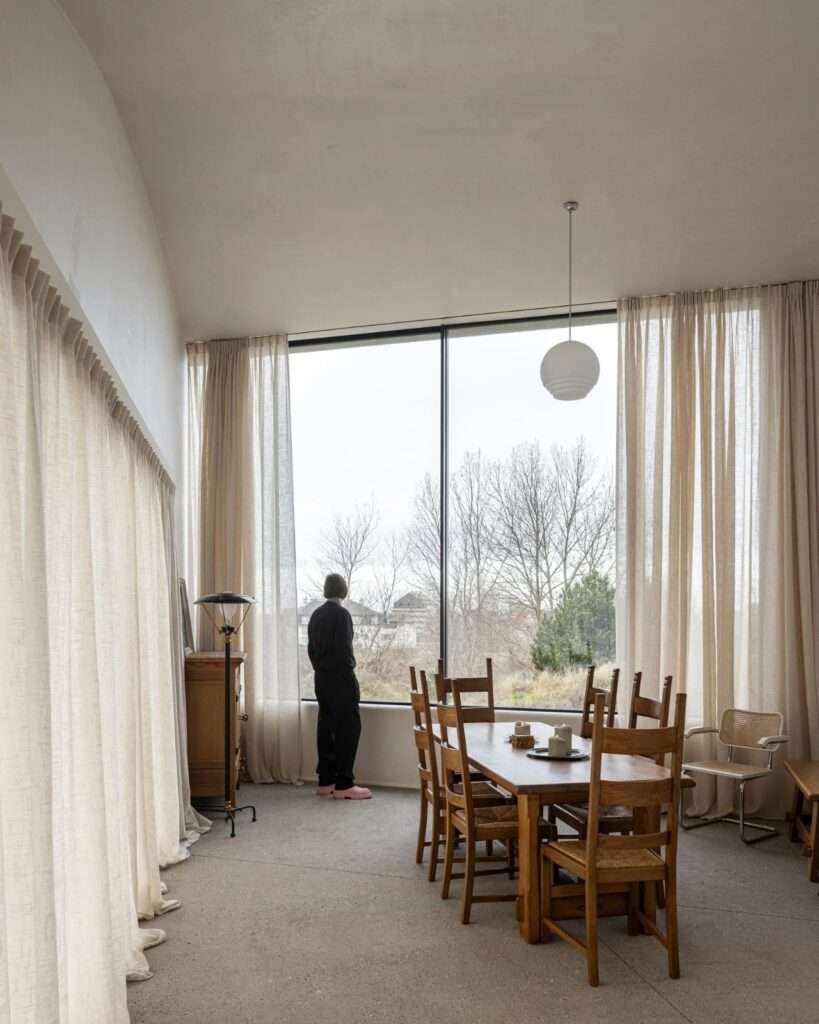 Modern dining room with large windows overlooking a coastal landscape, featuring wooden furniture and linen curtains.