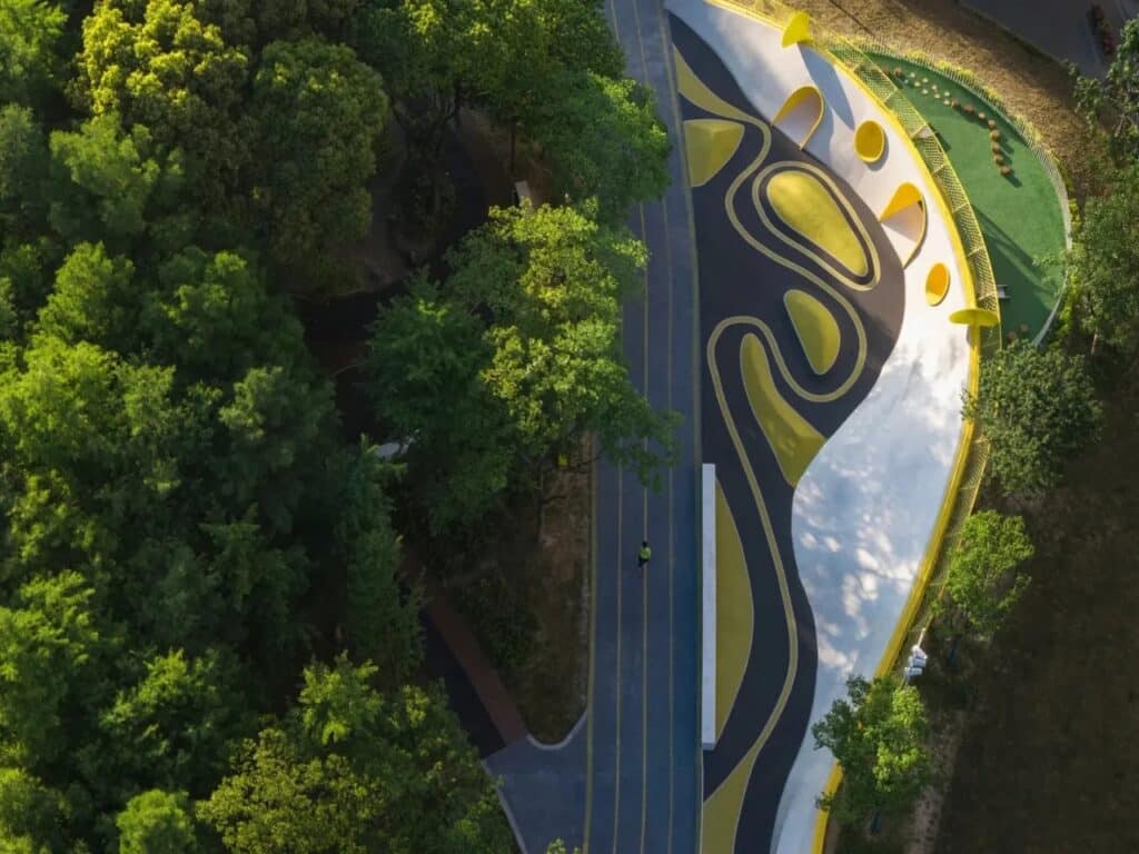 Top-down aerial view of a colorful playground with yellow, black, and grey wavy patterns next to a running track and forest.