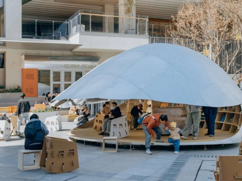 A daytime view of the "Book Shell" pavilion showing people and children interacting on the deck and using scattered movable cardboard chairs in the square.