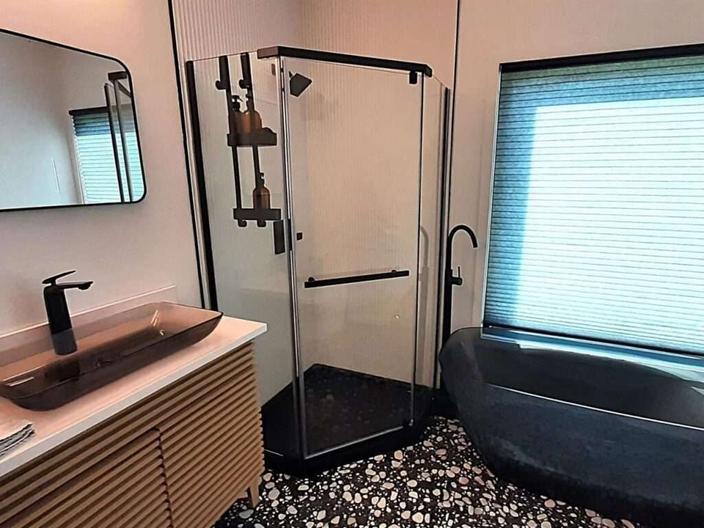 Modern bathroom interior in a container house featuring a glass shower, stone tub, and pebble-style flooring.